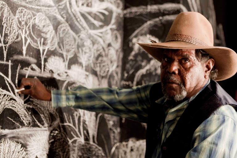 A middle-aged Aboriginal man in a hat and wearing a checked shirt and vest is drawing in chalk on a blackboard.
