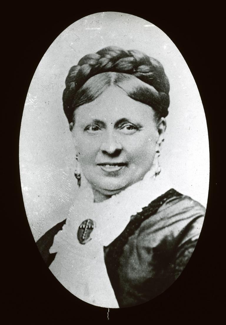 Glass slide of Emma Hardinge Britten : Black and white glass slide showing the head and shoulders portrait of a woman wearing a pale blouse with a brooch attached and a dark jacket, turning head to her left, looking left, smiling.