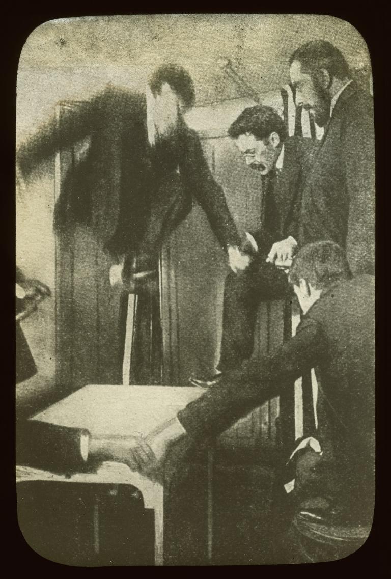 Séance glass slide Sepia glass slide showing approximately five men and a spirit joining hands around a table, looking down at the table.