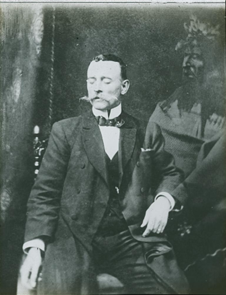 Glass slide of spirit Black and white glass slide showing a man sitting on a chair with his eyes closed and a spirit partly superimposed over his left arm and chest.