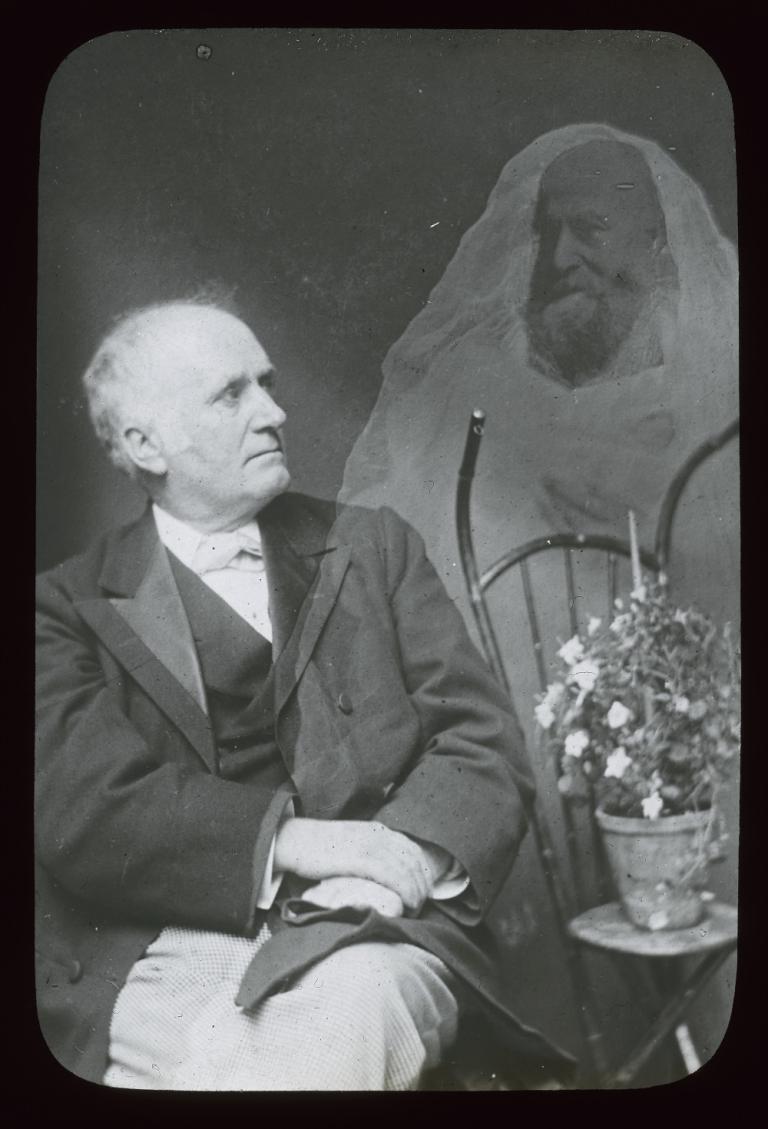 Man looking at a hovering spirit Black and white glass slide showing a man sitting on a chair with arms folded over his knees, looking to his left at a spirit, partially superimposed over his left arm and chest.