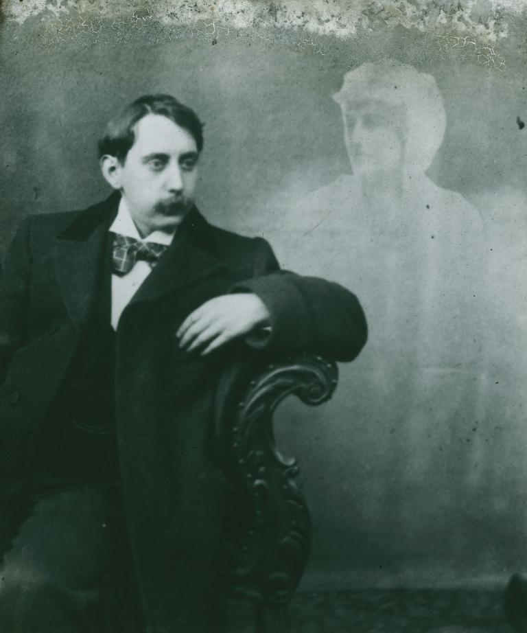 Fashionably dressed man with spirit Black and white glass slide showing a man sitting sideways on a chair, elbow resting over side of chair, with a spirit hovering at his left.