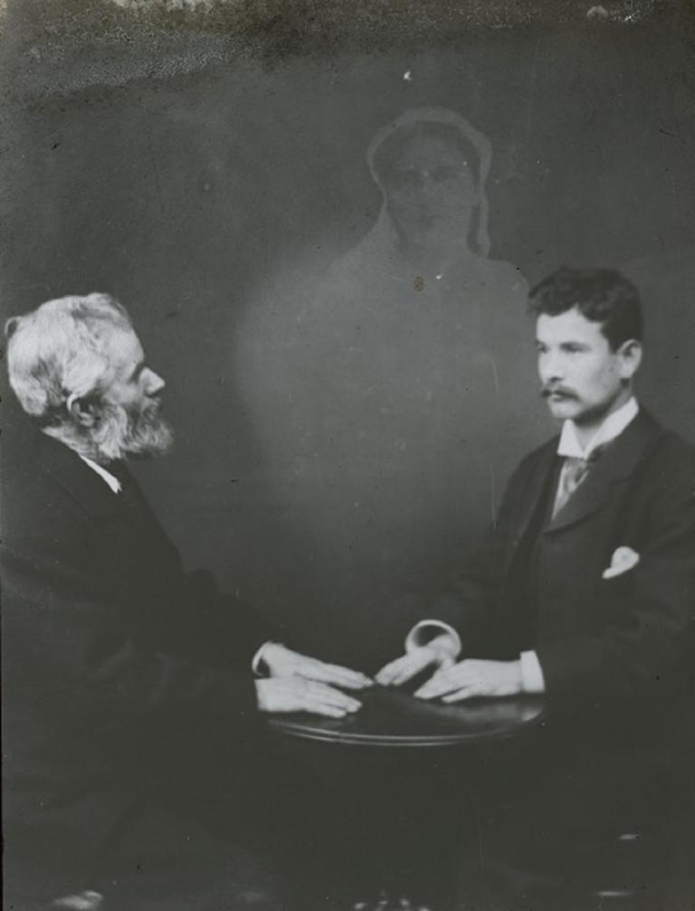 Two men conjure a spirit Black and white glass slide showing two men sitting opposite each other, hands resting on a table, a spirit hovering behind them.