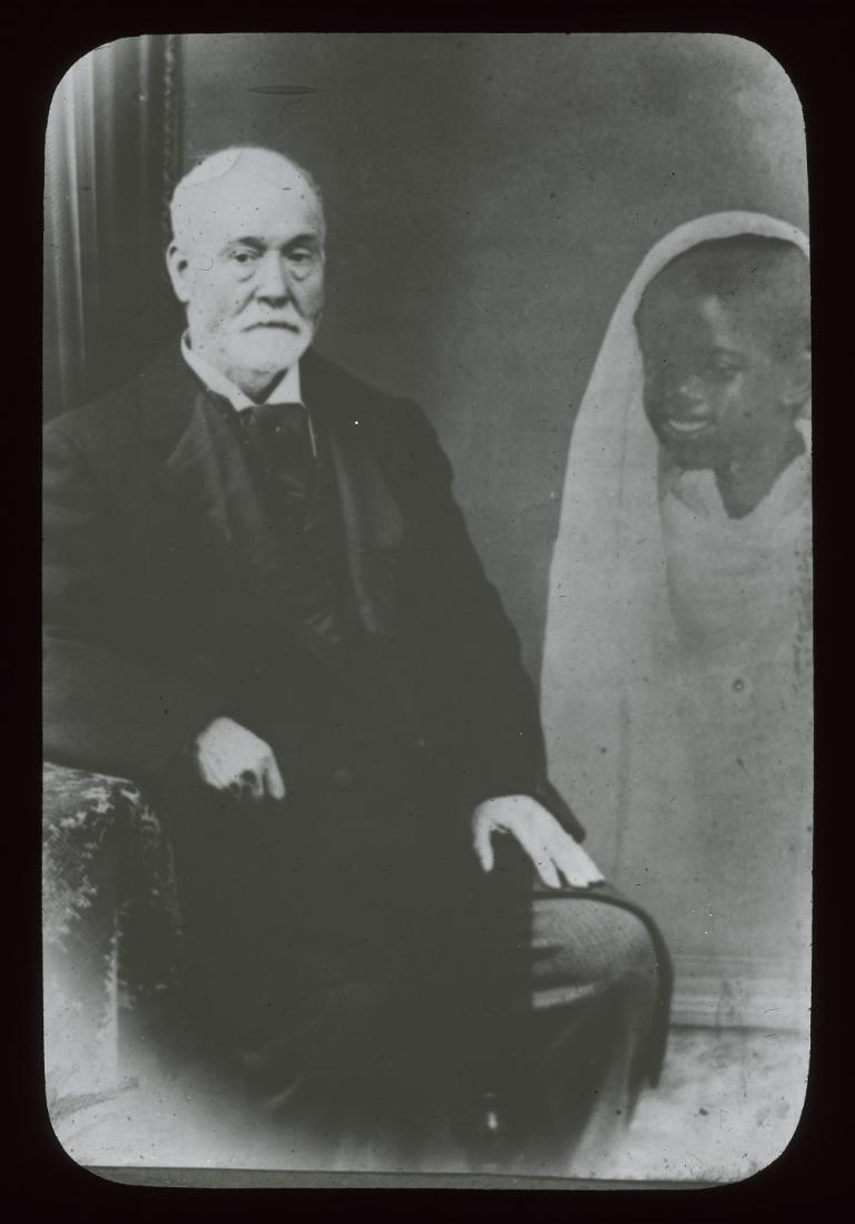 Glass slide of a spirit Black and white glass slide showing a man sitting on a chair, looking into camera, a spirit hovering at his left knee.