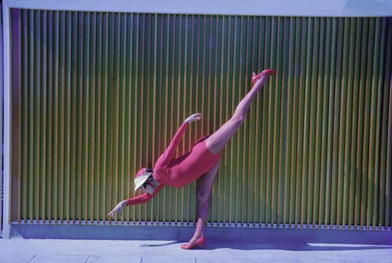 A colour photo of Annette Kellerman striking a dramatic pose. She is standing on one leg with her other leg in the air above her and her arms stretched out. She is dressed in pink.