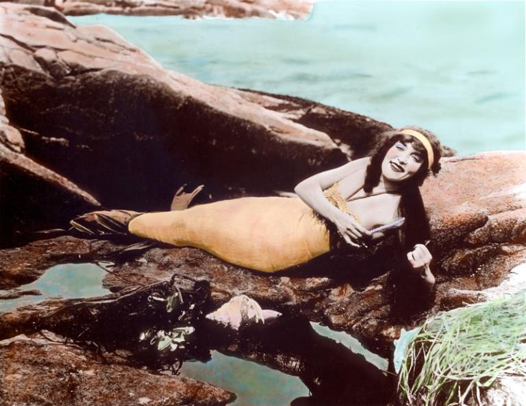 A hand-tinted photograph of Annette Kellerman wearing a mermaid costume and lying on rocks near the water.