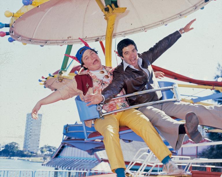 Wonder World! reporter Jonathan Coleman and Simon Townsend sitting on a fun park ride at Sydney's Luna Park. They are posing with their arms outstretched for the camera. 