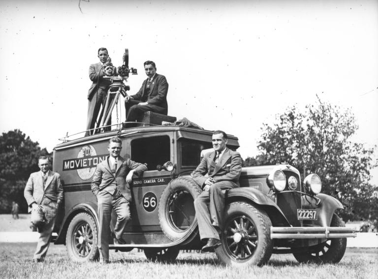 Bill Trerise with a camera on a tripod and one other man on top of Movietone Sound camera car 56, with three other men on the ground.