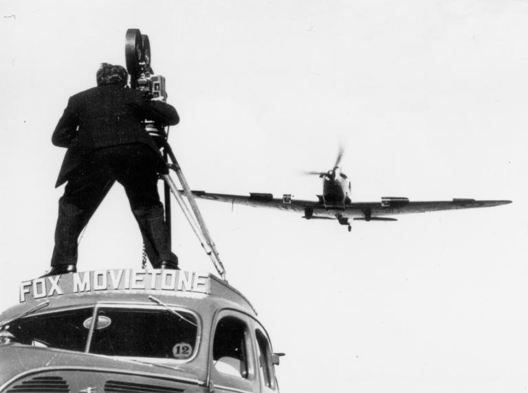 Fox Movietone cameraman standing on a car filming a fighter aircraft flying overhead