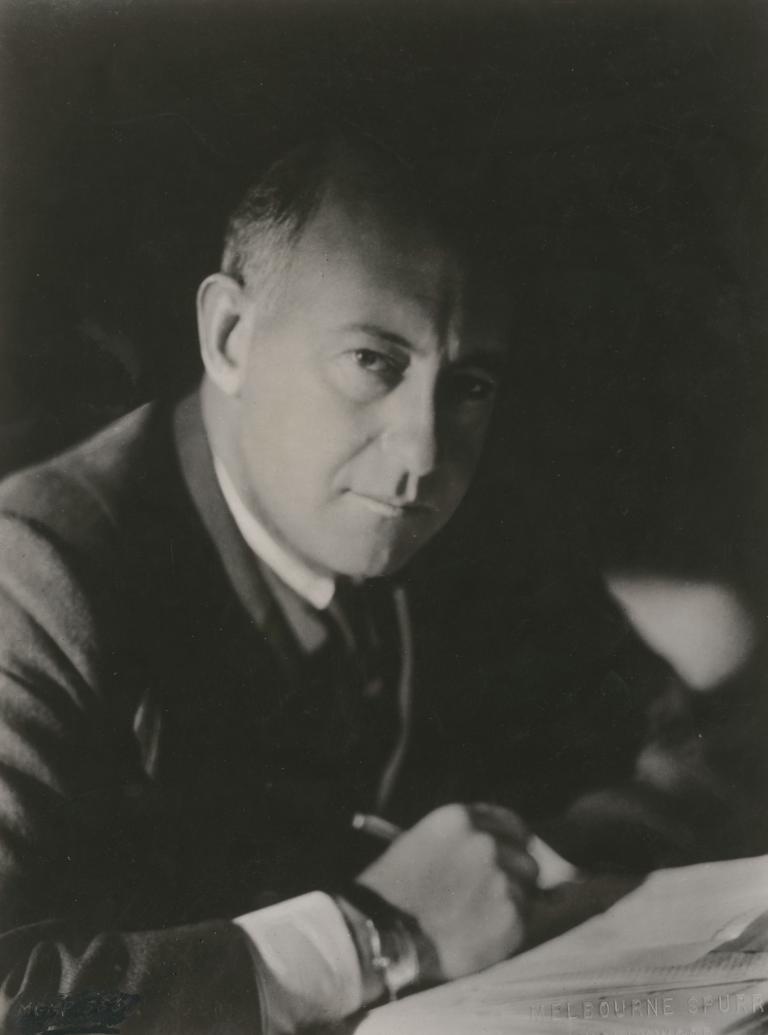 Cecil B DeMille seated at his desk and looking to camera, 1930.