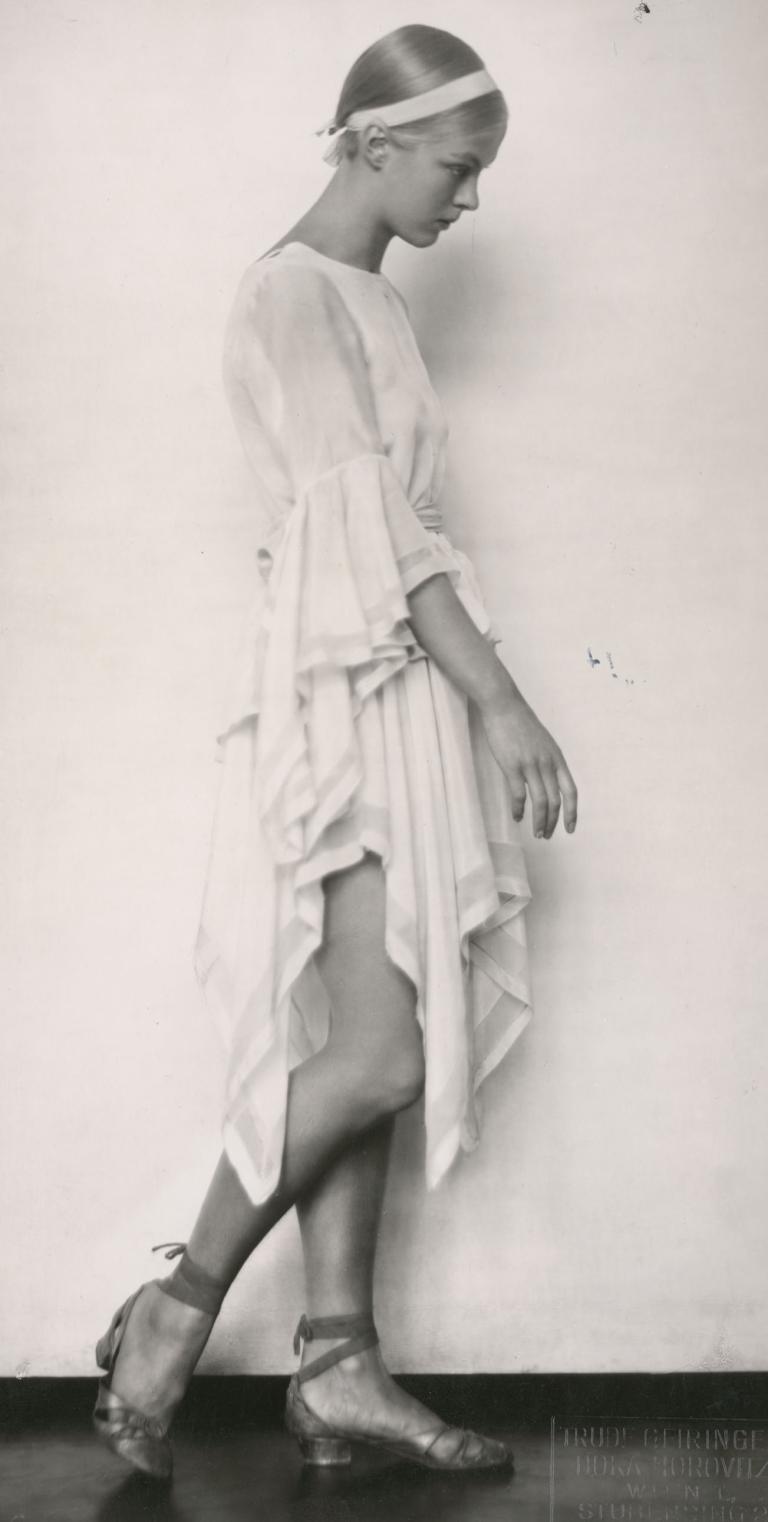 A young woman in a white dress and matching hair band stands side-on to the camera, looking downwards, in a still from the film Mädchen in Uniform, 1931.
