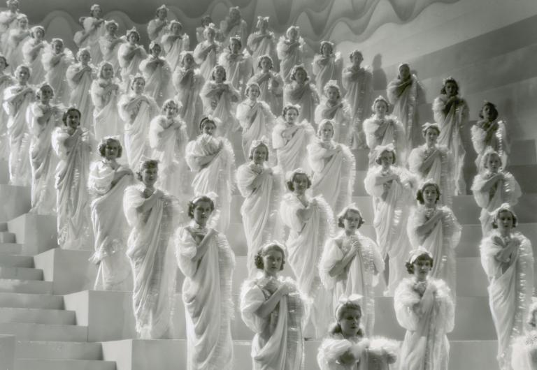 Women in evening dresses stand in diagonal rows on a staircase in a scene from the Austrian film Premiere, 1937.