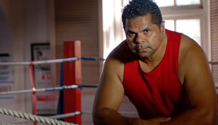 A wrestler in a red singlet leans on the rope of wrestling ring