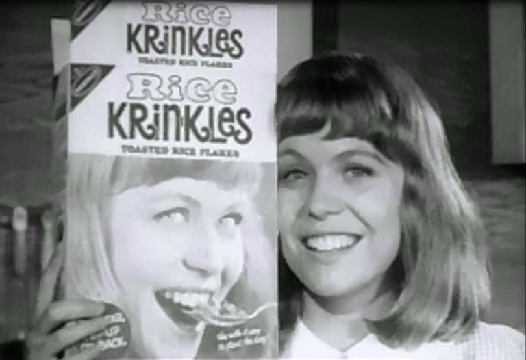 Jacki Weaver, smiling and holding a box of 'Rice Krinkles', featuring her face on the front. In the box, Jacqui is seen eating the product.