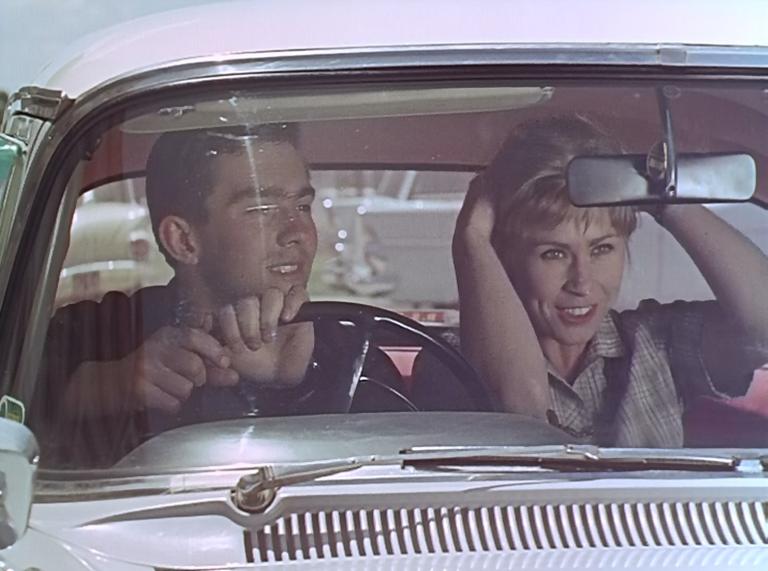 A couple in a 1960s car. The man is driving, as his female companion uses the rear-view mirror to fix her hair