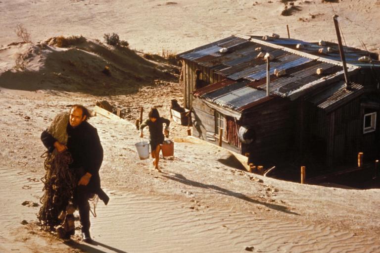 Peter Cummins as Hideaway Tom walking up the sand dunes with fishing nets slung over his shoulder and Greg Rowe as Storm Boy following him the dunes carrying two pails of water. Their simple corrugated iron humpy is below them.