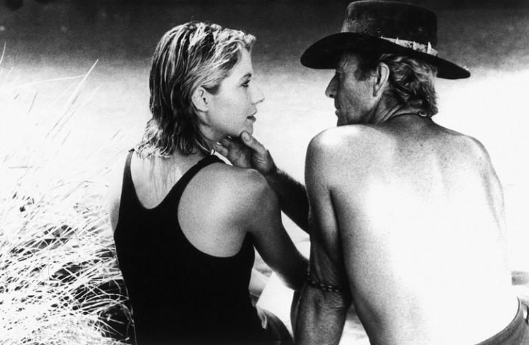 Linda Kozlowski as Sue Charlton and Paul Hogan as Mick ‘Crocodile’ Dundee sitting by a waterhole. He is touching her face and they are staring at each other.