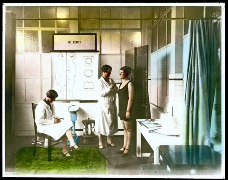 A woman wearing a white coat measures another woman in her underwear. A third woman (also in a white coat) takes notes. They are in a medical-looking room with a sign that says 'BE EXACT!'.