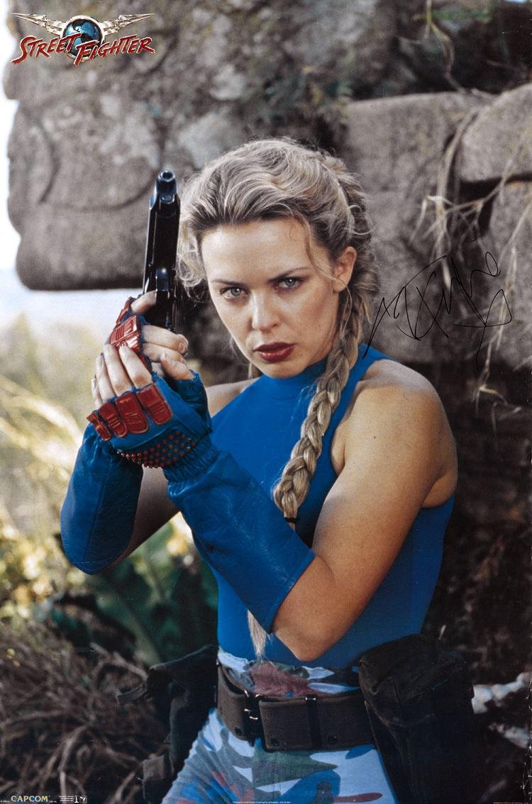 Kylie Minogue poses holding a gun wearing a blue camouflage costume and fingerless gloves. Her steely gaze directed to the camera.