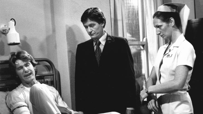 Dr Brian Denham (Michael Beecher) and Sister Scott (Cornelia Frances) stand by the the bed of patient, Craig Rothwell (John Walbon).