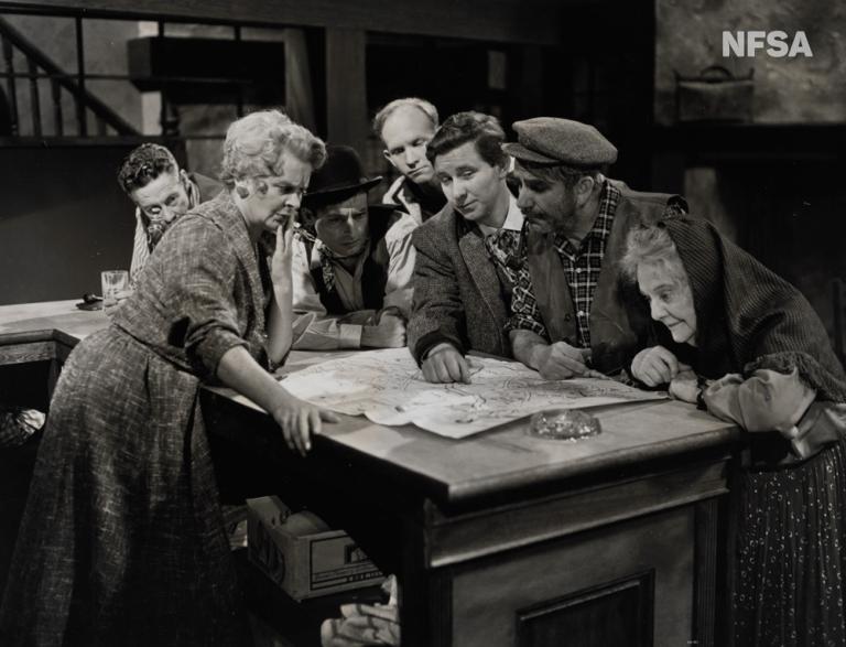 A group of people are crowded around a table in a still from a television production.