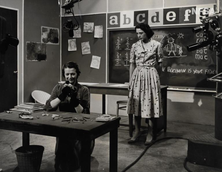 A production still of two women in a school room setting. A boom is visible in the background.
