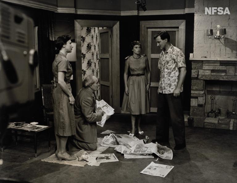 Three people in a studio set feature in this black and white production still.