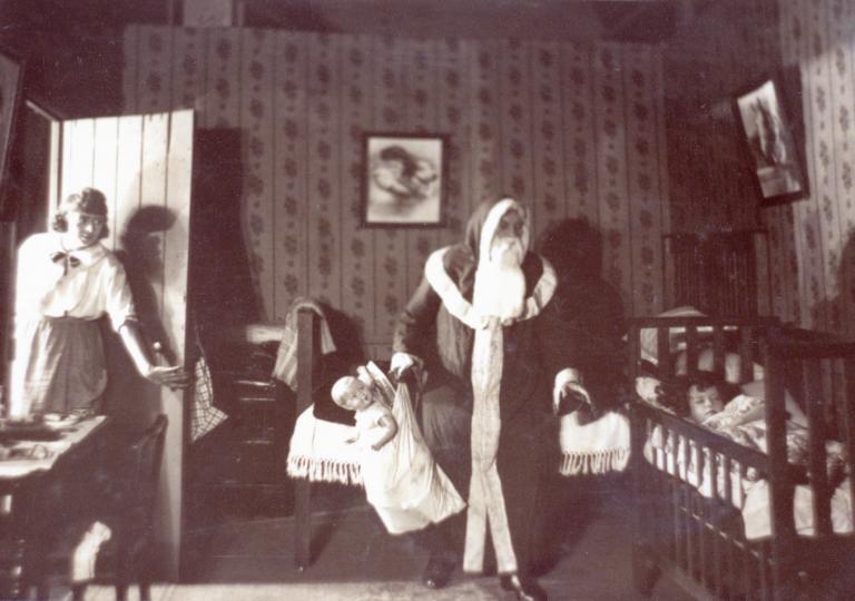 Actor Arthur Tauchert, dressed as Santa Claus and holding a large doll, looks at a sleeping child. Lottie Lyell watches from behind the door.
