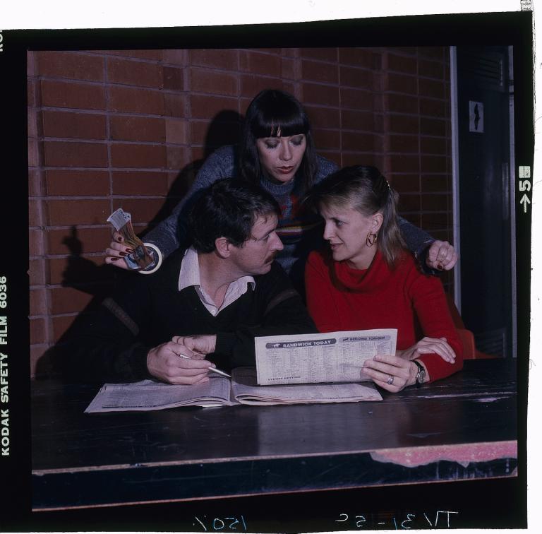 Actress Sandy Gore holding a couple of $20 notes, stands over her colleagues Gerard Maguire and Jane Clifton, who are looking at each other. Clifton is holding the newspaper, which reads 'Randwick Today'.