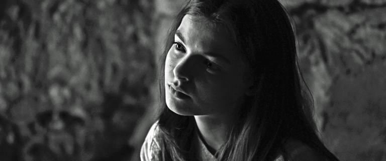 Still from the film Brolga Black and white, head and shoulders shot of young woman with a serious look on her face and head tilted to the side. Still from the film Brolga.