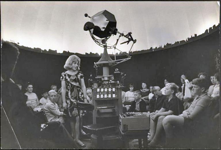 A woman stands in the middle of a crowd of young kids at an observatory