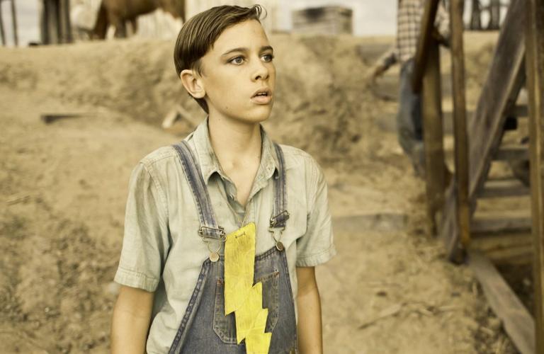 A young boy wearing a short-sleeved collared shirt, a pair of denim overalls and a large yellow lightening bolt pinned to the front of his shirt.
