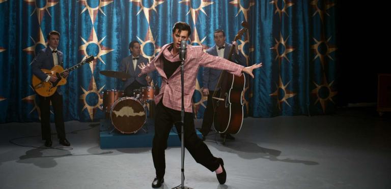 Austin Butler as Elvis Presely singing with his 4-piece band on stage in a recreation of a 1950s TV studio. Elvis is wearing a pink blazer and the group are performing in front of a turqouise curtain with sun symbols on it