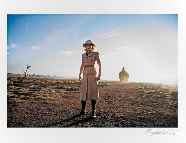 Actor Nicole Kidman wearing a safari-style outfit with a long skirt, belted shirt and knee-high boots and a hat. She is standing in a dusty outback setting during the filming of Baz Luhrmann's film Australia.  The photo has been signed in the bottom right corner by photographer Douglas Kirkland.