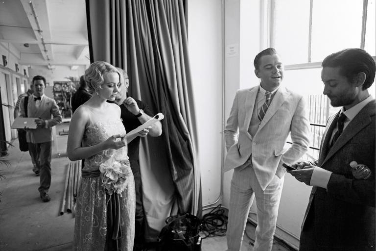 Actors Carey Mulligan, Leonardo DiCaprio and Tobey Maguire standing in a room in a semi-circle. They are on a film set dressed in 1920s costumes.