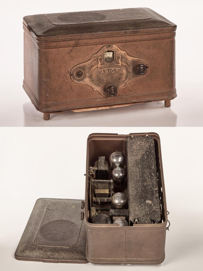 An electric radio from the late 1920s in a metal box with two dials on the front and a removable lid. 