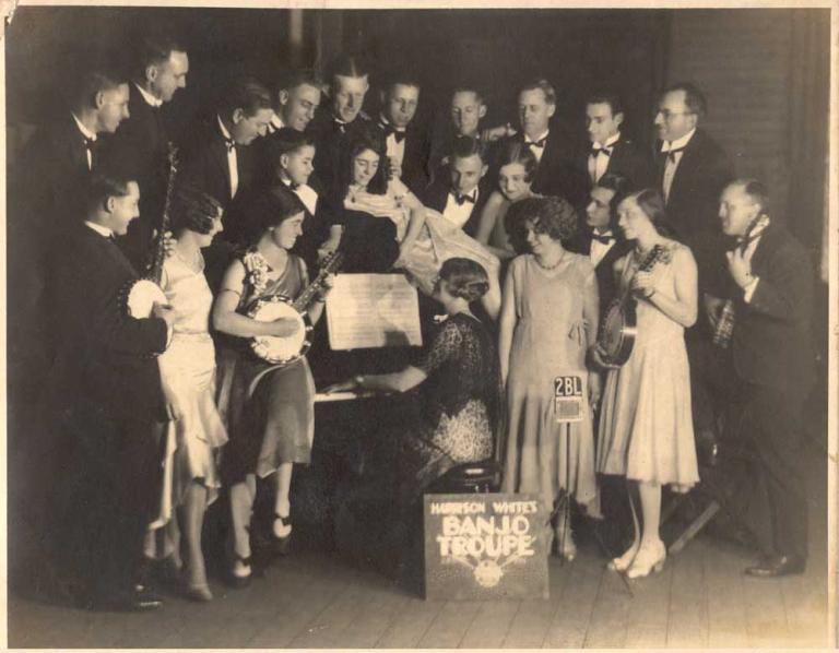 A group of people holding banjos and standing around a piano.