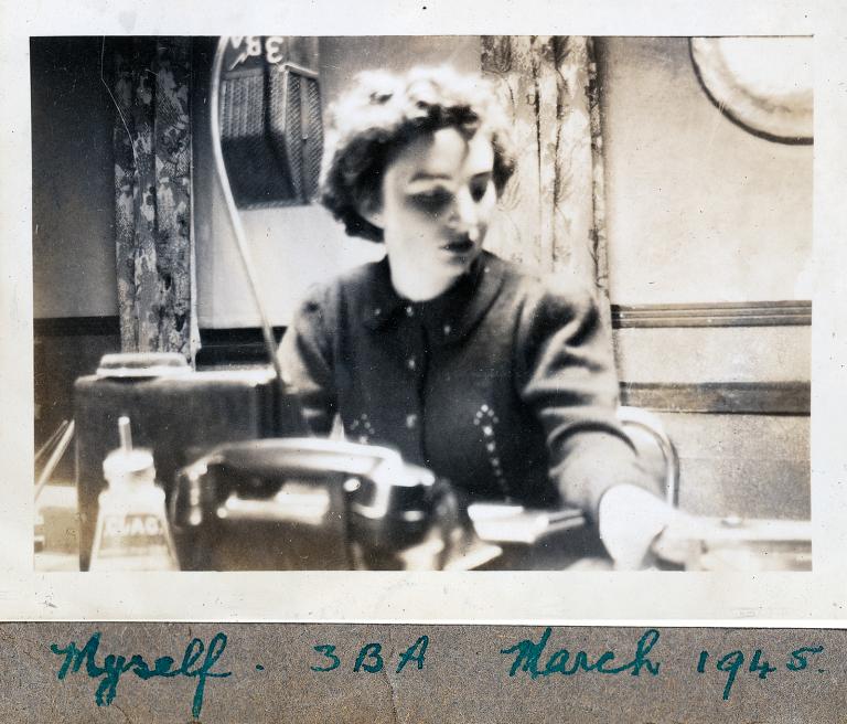 A woman sitting at a desk. She is looking at something on the desk and there is a telephone and a radio microphone in front of her. Below the photo is handwriting that says 'Myself. 3BA. March 1945'