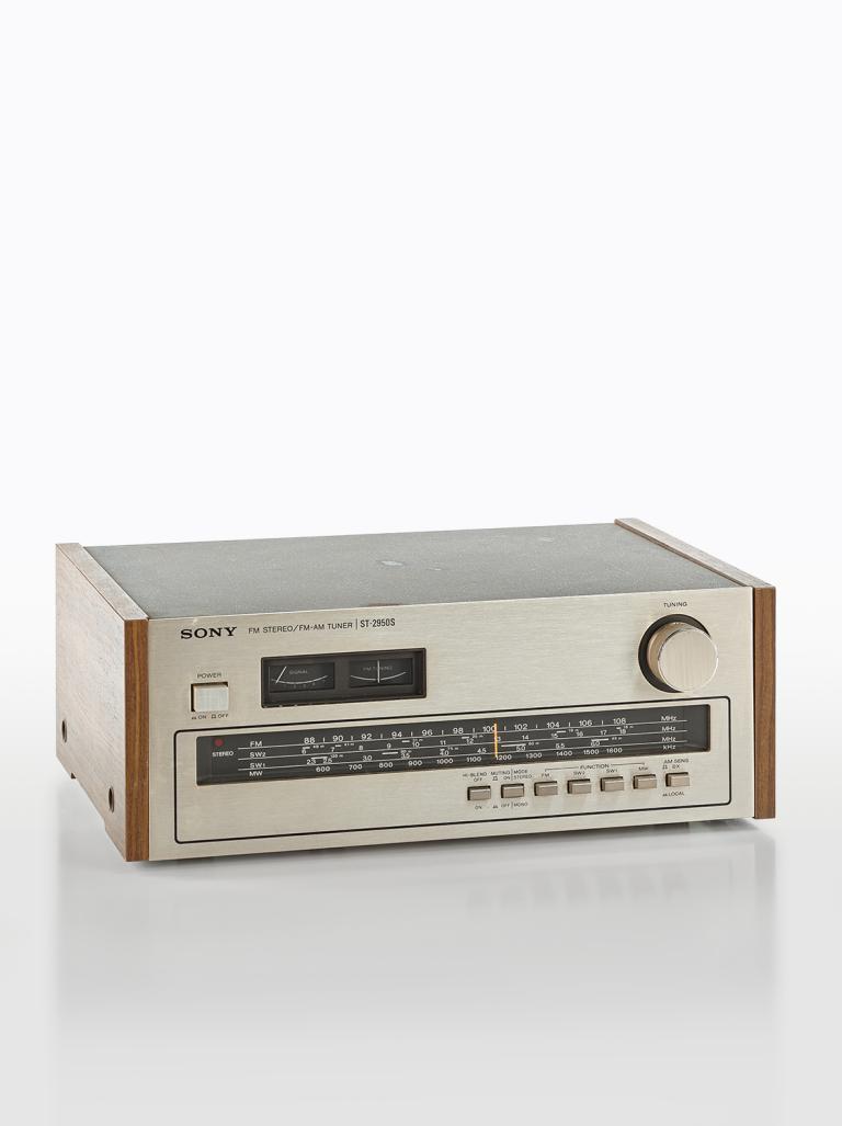 A Sony FM radio tuner from the 1970s. It is a rectangular silver shape with a tuning dial and radio bands listed on the front in rows.
