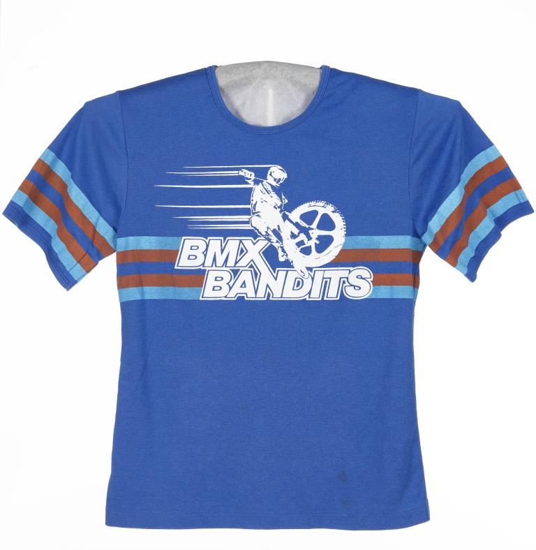 A photo of a blue t-shirt with the text BMX Bandits on it. There's a white logo with a driver and a wheel racing on top of the text. 