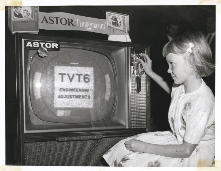 A small girl is on the right hand side of the photo. She is looking towards the tv screen which has the holding title "TVT6" on it. The photo is in Black and White