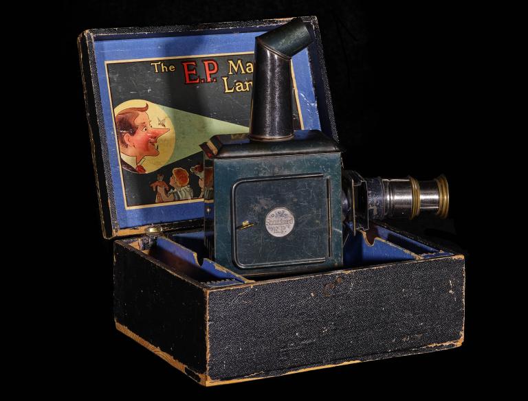 A photo of a lantern projector in its black wooden box. The projector is also black with two tubes coming from it, one on top, the other on the side.