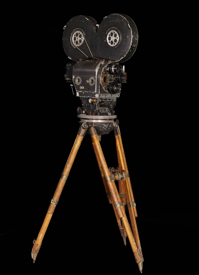 A photo of a black camera with two reels sitting atop a light wood tripod.