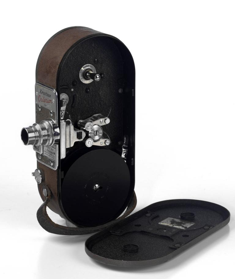 A photo of a movie camera that is brown in colour with a bright silver lens at the front. The lid is off and to the side allowing you to see the reels within the case.