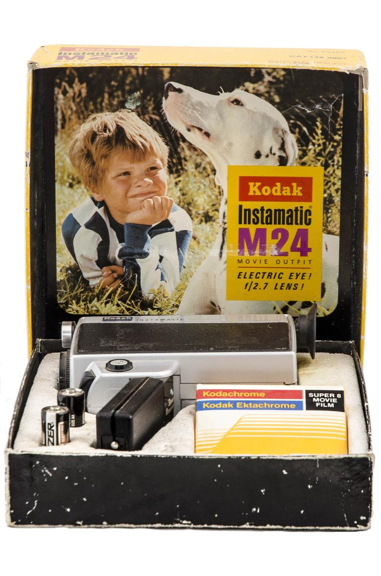 A photo of a home movie camera in its box. The handheld camera is black and grey in design and the box it's in has a picture of a small box next to a dog. The Kodak brand logo and model "Instamatic M24" is visible. 