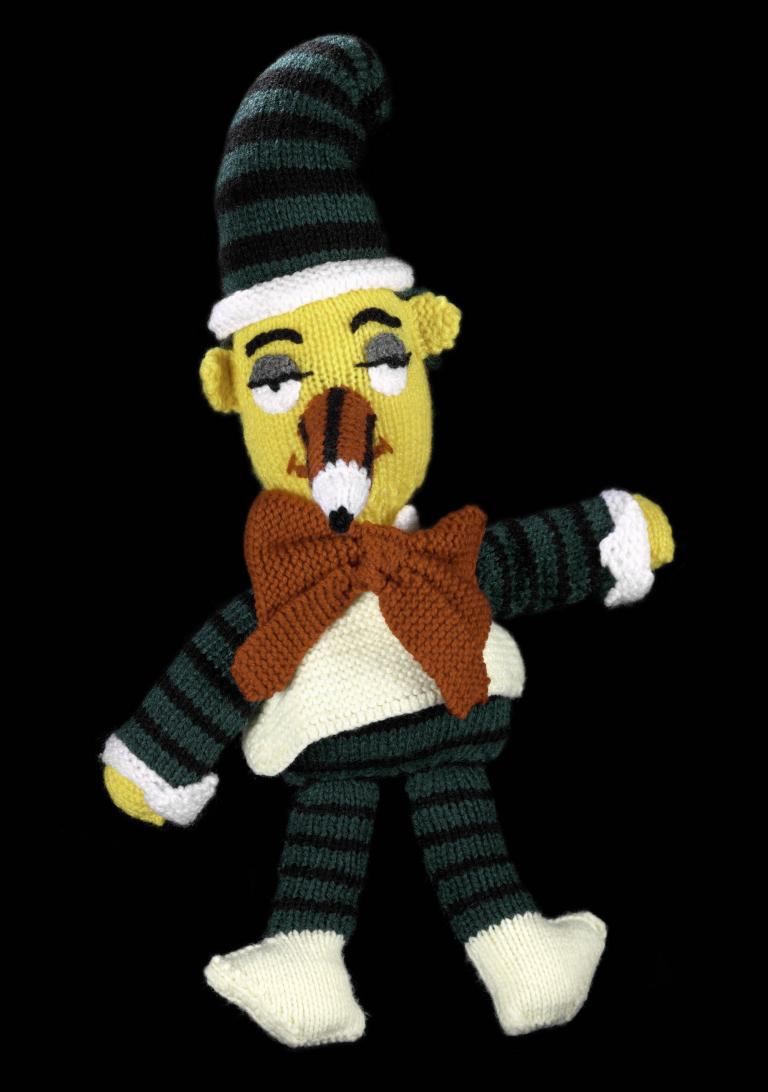 A photo of a knitted plush toy of Mr Squiggle. They're wearing dark blue and black stropped clothing, an oversized red bowtie and white shoes. 
