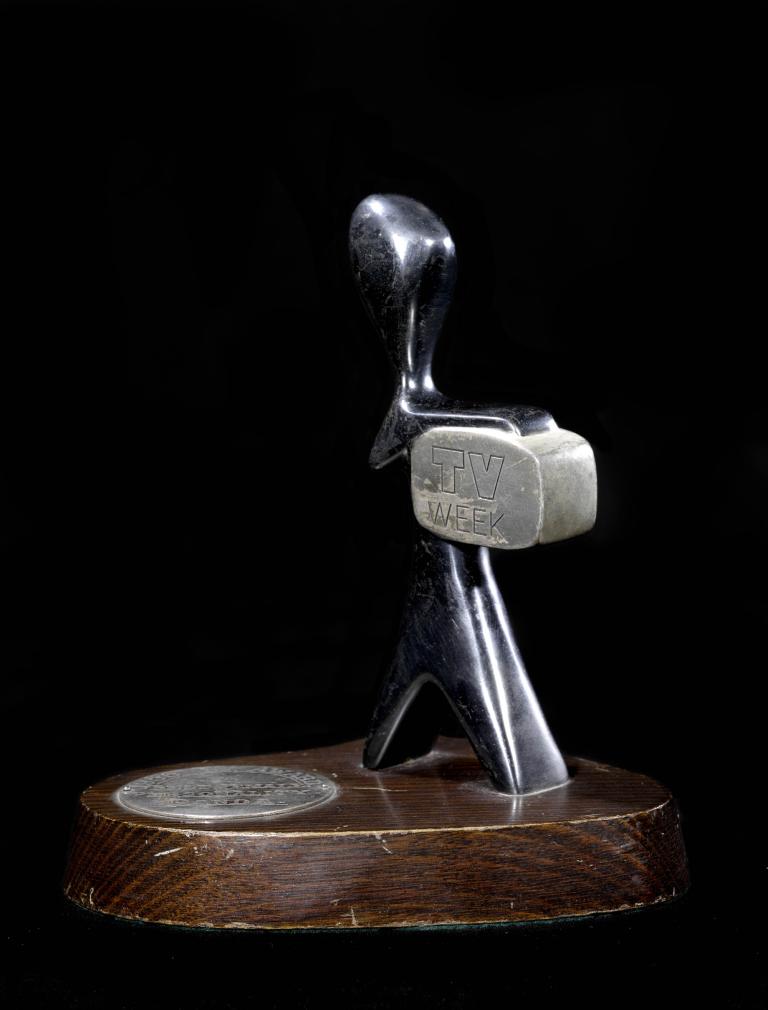 A photo of a TV week award. It is on a wooden base with an abstract silver sculpture representing a person holding a tv screen.