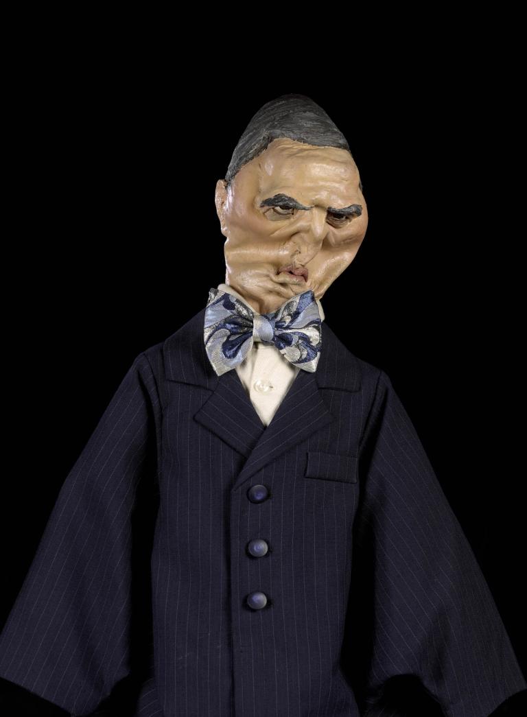 A photograph of a puppet wearing a black suit and a blue patterned bow tie. Its face is weathered.