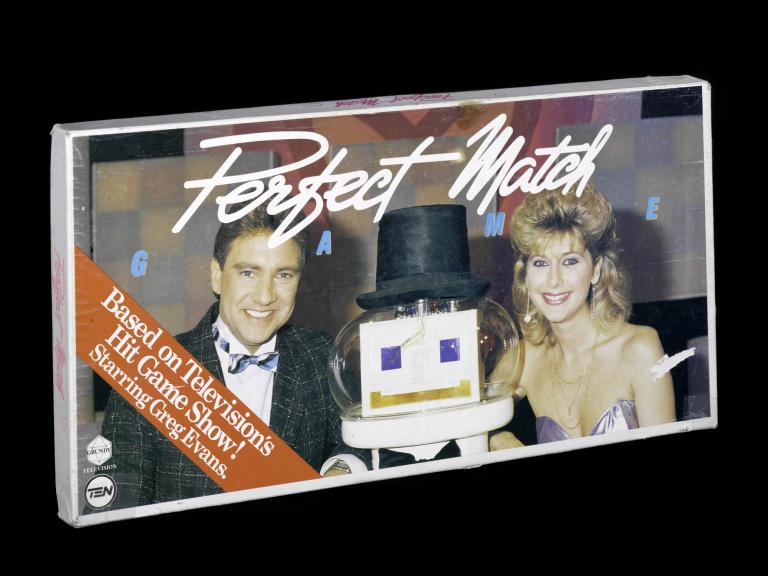 A photo of the Perfect Match boardgame on a black background. A man and woman are picture on the front with a robot wearing a top hat sat between them. 