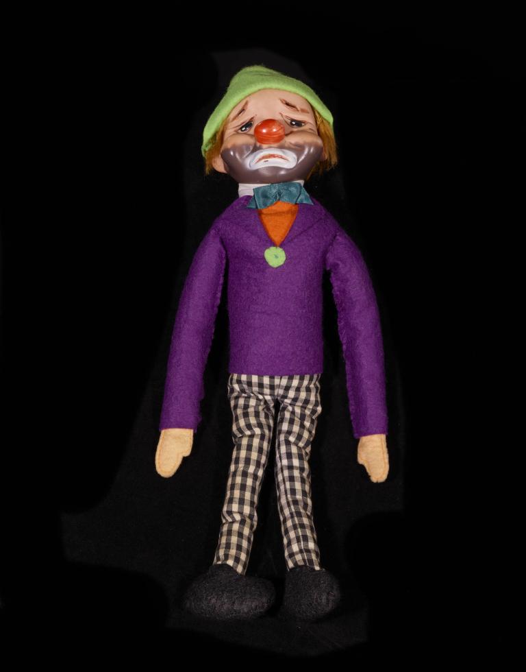 A photo of a doll with a purple top, green hat and plaid brown trousers.